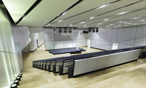 ferrum Blick von der obersten Reihe des Konferenzraums ferrum in den hell erleuchteten Saal. Man erkennt die absteigenden Sitzreihen, die wie ein Hörsaal aufgebaut sind. Vor den Sitzreihen befindet sich, mit etwas Abstand, ein niedriges Podest mit einem Rednerpult. Links vom Podest befindet sich ein Durchgang zum Foyer in der dritten Ebene.