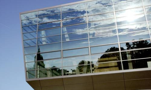 Bild: darmstadtium Blick auf die Ecke der Glasfassade des darmstadtiums, die aus vielen kleineren Fenstern besteht. Man erkennt im inneren des Gebäudes Konferenzräume über zwei Ebenen, in denen sich Betonpfeiler befinden. In der Glasfront spiegelt sich der Turm des Hessischen Landesmuseums.