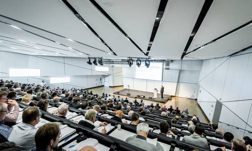 ferrum, Copyright: Fotografie Schepp Blick von den oberen Reihen hinunter in den Konferenzraum ferrum. Die Teilnehmer sitzen während einer Tagung in ansteigenden Stuhlreihen, der Saal ist vollbesetzt. Die Tische vor ihren Sitzen sind ausgeklappt. Die Teilnehmer hören dem Redner zu, der sich in der Bildmitte befindet.
