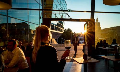 ©Gaul Catering GmbH & Co Blick aus "CALLA die weinbar" in die untergehende Sonne über dem Hessischen Landesmuseum. Gäste sitzen an den Tischen und vor der Tür. Ein Gast kommt die geöffnete Tür hinein, eine Dame scheint mit einem Glas Wein auf jemanden zu warten.