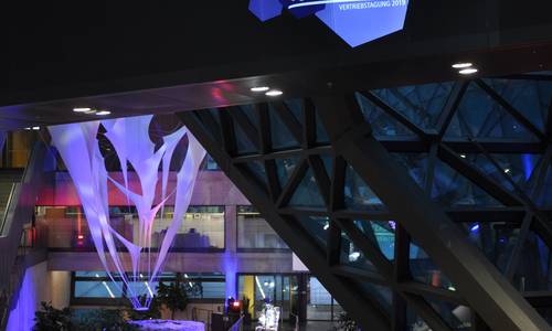 Calla und Foyer 0.02 Copyright conceptx Blick von der Haupttreppe hinunter zum Haupteingang. Im atrium sind Stände aufgebaut. Die Zisterne des Lichtes und die Betonpfeiler sind blau angeleuchtet.