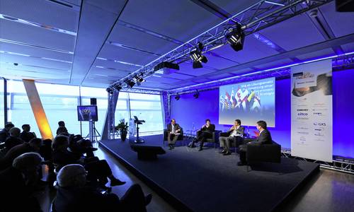 Konferenzraum Während einer Veranstaltung diskutieren vier Personen in einem Konferenzraum auf einem in blau angeleuchteten Podium . Im Vordergrund links sitzen die Teilnehmer der Veranstaltung im Dunkeln und hören zu.