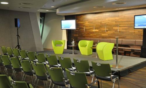 Lounge Vor der holzvertäfelten, angeleuchteten Wand stehen drei grüne Sessel auf einem niedrigem Podium. Vor dem Podium befinden sich Stuhlreihen.
