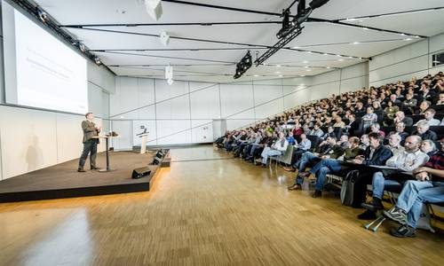 ferrum, Copyright: Fotografie Schepp Blick von der linken Seite durch den Konferenzraum ferrum. Die Teilnehmer sitzen während einer Tagung in ansteigenden Stuhlreihen. Der Saal ist vollbesetzt, die Schreibtische vor ihren Sitzen sind ausgeklappt. Die Teilnehmer hören dem Redner zu, der sich am linken Bildrand auf einem niedrigen Podest befindet.