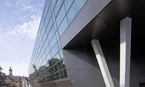 Bild: darmstadtium Seitliche Ansicht der Glasfassade von der Erich-Ollenhauer-Promenade aus. Zwei Betonpfeiler befinden sich am unteren Bildrand. Im Bildhintergrund befindet sich das Residenzschloss.