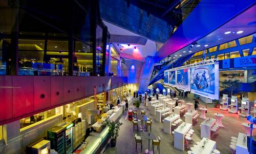Foyer 0.02 atrium Einblick in eine Veranstaltung im darmstadtium. Im atrium wurde zur Seite des Restaurants Calla eine Cateringfläche mit Stehtischen aufgebaut. Zur Seite der Stadtmauer befinden sich Stände von Ausstellern. Das Licht im atrium ist in blau und rot gehalten.