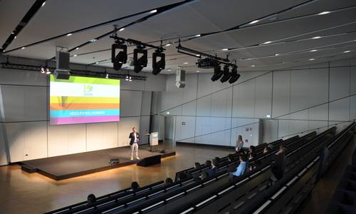 ferrum Seitlicher Blick in den Konferenzraum ferrum auf die Bühne. Einige Personen sitzen in den ansteigenden Reihen des Raumes. Die bunte Präsentation auf der Leinwand leuchtet intensiv, ein Redner befindet sich auf der Bühne.