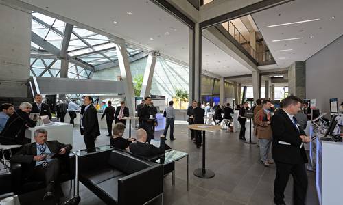 Foyer 1.05 Vor dem großen Saal spectrum befinden sich Teilnehmer einer Veranstaltung. Sie sitzen links bei den aufgebauten Sitzgelegenheiten, stehen an Stehtischen oder an einem Schalter. Auf der rechten Seite befindet sich der große Saal spectrum. In der Bildmitte führt die Haupttreppe zum atrium.