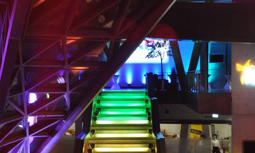 Foyer 0.02 atrium Im atrium des darmstadtiums findet eine Abendveranstaltung statt. Die Haupttreppe ist in den Farben grün, gelb und orange, das Foyer am oberen Ende der Treppe in blau, die Betonstreben in lila angeleuchtet. Im unteren Bereich des Bildes befinden sich die Teilnehmer.