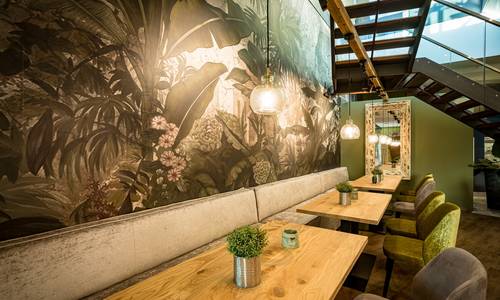 ©Gaul Catering GmbH & Co An einer Wand befinden sich eine Sitzgelegenheit im Restaurant CALLA. Direkt an der mit Pflanzen bemalten Wand befindet sich eine durchgängige Sitzbank drei kleinere Tische und Stühle befinden sich auf der gegenüberliegenden Seite. Über den Tischen hängen erleuchtete Lampen. Am oberen Bildrand erkennt man die Treppe, die in die Weinbar führt.
