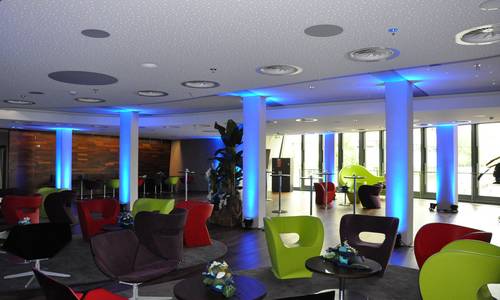 Lounge In der lounge des darmstadtiums sind grüne, weinrote und rote Sessel unterschiedlichstem Design arrangiert. Auf runden, grauen Teppichen stehen die Sesselgruppen um kleine Beistelltischen herum. In der Bildmitte stehen Stehtische, an der beleuchteten linken Wand und am gegenüberliegenden Fenster befinden sich weitere Sitzgelegenheiten. Die Stützpfeiler in der Raummitte sind blau angestrahlt.
