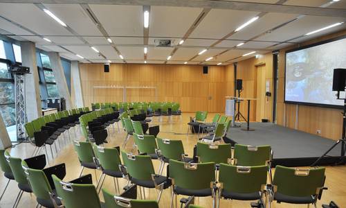 3.089 helium 2 Blick in den Konferenzraum helium2, einer Raumkombination der Konferenzräume helium und neon. Auf der linken Bildseite sind vor der Fensterfront, Stühle in drei kleineren Blöcke zu einem Halbkreis aufgebaut. Auf der gegenüberliegenden Seite befindet sich ein Podium und eine Leinwand. Fünf Moderatorenplätze stehen vor dem Podium und blicken zu den Stuhlreihen. Neben dem Podium befinden sich Lautsprecher und eine Leinwand, auf dem Podium zwei Rednerpulte.