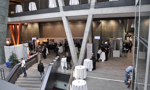 Foyer 1.05 Im Foyer 1.05 sind Messestände aufgebaut. Interessierte Kunden befinden sie an den Ständen. Am linken Bildrand befindet sich die Haupttreppe, die ins atrium führt.