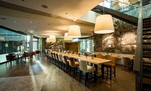 ©Gaul Catering GmbH & Co Blick in das Restaurant CALLA, wo mitten im Raum eine lange Tafel aufgebaut ist, darüber hängen große Leuchten. An der linken Seite, der Seite zum atrium des darmstadtiums, stehen runde Tische mit Stühlen. Ebenso an der rechten Seite. Am äußersten rechten Rand erkennt man die Treppe, die zur Weinbar führt.