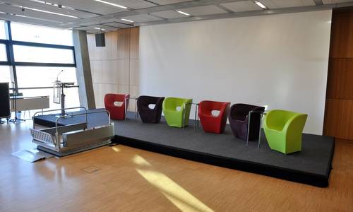 3.023 germanium 2, Bühne mit barrierefreiem Zugang Blick in den Konferenzraum germanium2. Es befinden sich Sessel vor einer Leinwand auf einem niedrigen Podium. Über einen mobilen Rollstuhllift erreichen Rollstuhlfahrer das Podium. Am linken Rand des Podiums befindet sich ein Rednerpult.