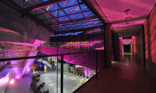 Blick ins atrium von der Ebene 2 Blick in den Flur der Konferenzebene 2 auf Höhe der Brücke, die zum Konferenzraum ferrum führt. Der Gang ist pink erleuchtet. Am linken Bildrand erkennt man das fast leere atrium, wo sich schwarze Sessel und Couch ungeordnet stehen.