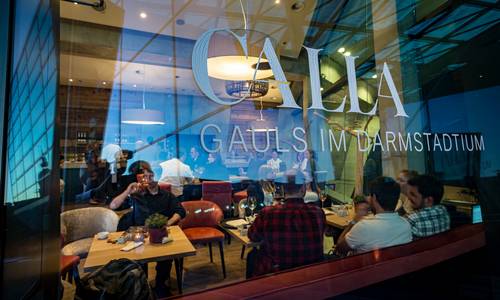 ©Gaul Catering GmbH & Co Blick durch die Glasfassade in die Weinbar des Restaurants CALLA. An den Tischen sitzen einige Gäste und unterhalten sich.