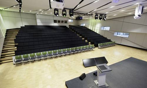 ferrum Blick von der Bühne auf den leeren Konferenzsaal „ferrum“. Im Vordergrund steht das Rednerpult auf einer niedrigen Bühne. Gegenüber befindet sich die ansteigende Sitzreihe, die an einen Hörsaal erinnert.