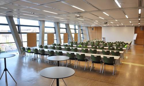 Dreierkombi 3.0234 Blick von der Tür in den Konferenzraum europiumum3, einer Raumkombination der Konferenzräume europium, germanium und hassium. Im Eingangsbereich befinden sich einige Stehtische. im Raum befinden sich Stühle an Tischreihen. Zwischen den Betonpfeilern, entlang der Fenster, stehen Pinnwände. Im Bildhintergrund ist ein Rednerpult und die Leinwand bereit gestellt worden.