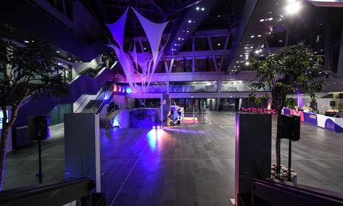 Foyer 0.02 atrium Copyright: Merck Blick von der Haupttreppe in Richtung Haupteingang. Im hell erleuchteten atrium sind auf der rechten und linken Raumseite Tische für eine Veranstaltung aufgebaut. Fast mittig auf dem Bild erkennt man die Zisterne des Lichtes und die Stadtmauer. Beides ist violett angeleuchtet.