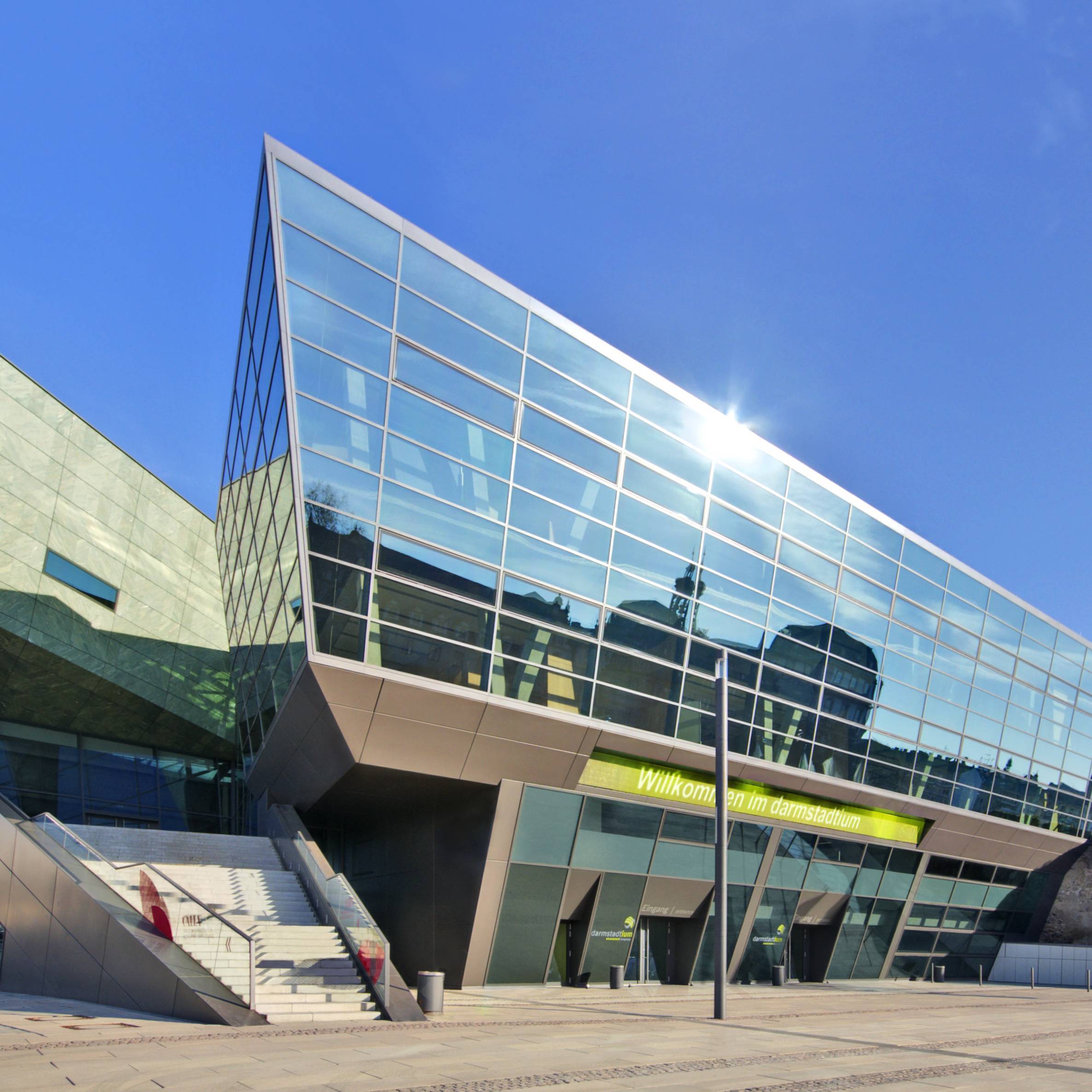 Nord-West-Ansicht, Schloßgraben Ecke Alexanderstraße, Copyright: darmstadtium Der Haupteingang und Vorplatz des darmstadtiums wurden bei strahlend blauem Himmel von der linken Seite aus fotografiert. Über dem Haupteingang spiegelt sich das Residenzschloss, das sich auf der anderen Straßenseite befindet.